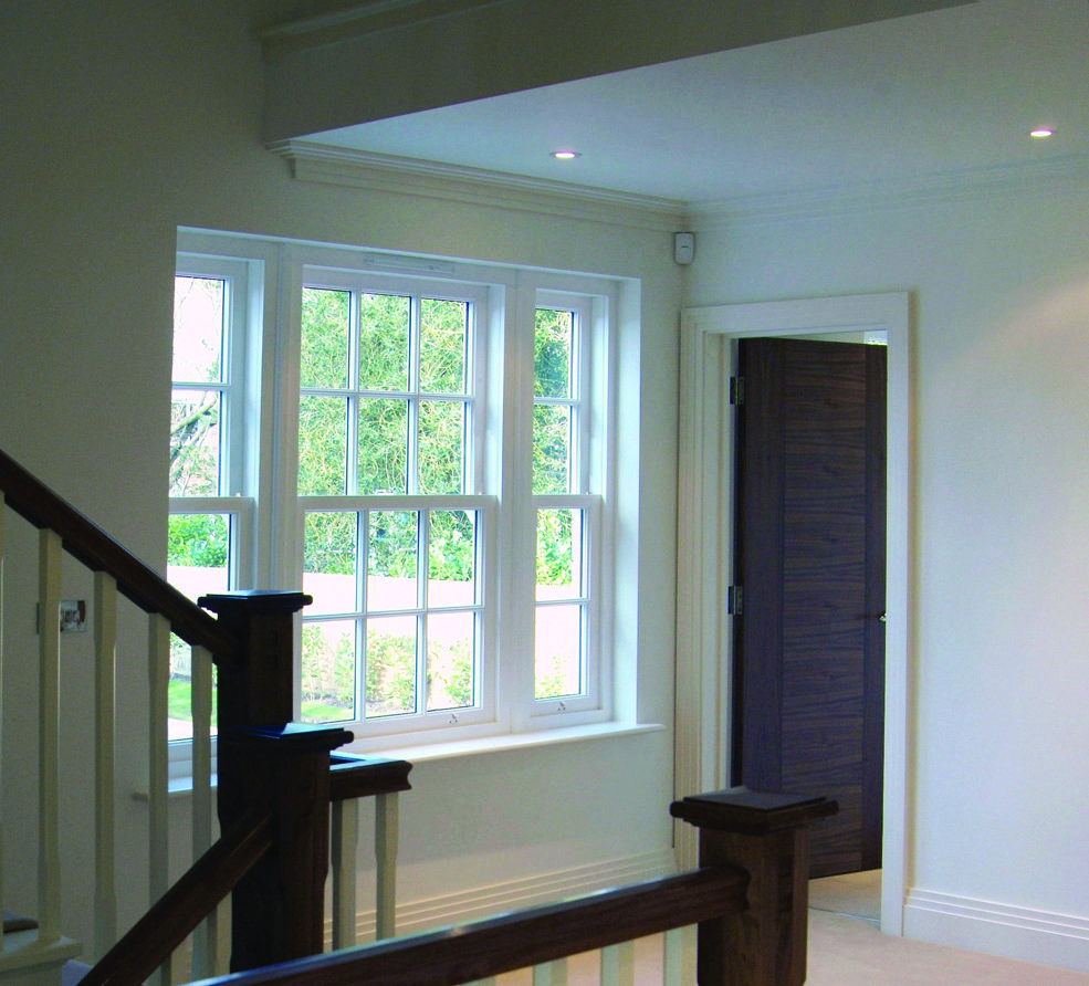A bright hallway with white walls, large windows, a wooden staircase, and a dark wood door.