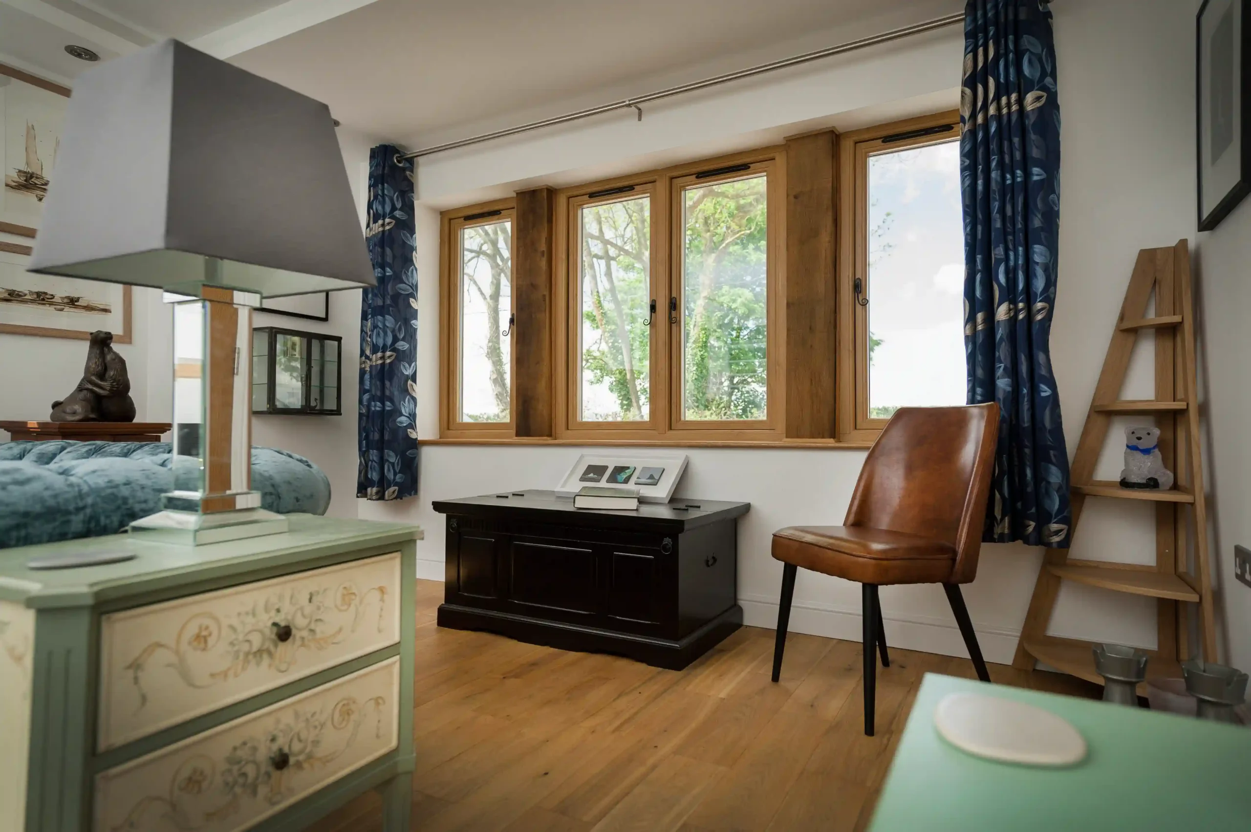 A bright bedroom featuring a large window with wooden frames, blue patterned curtains, and a brown leather chair.