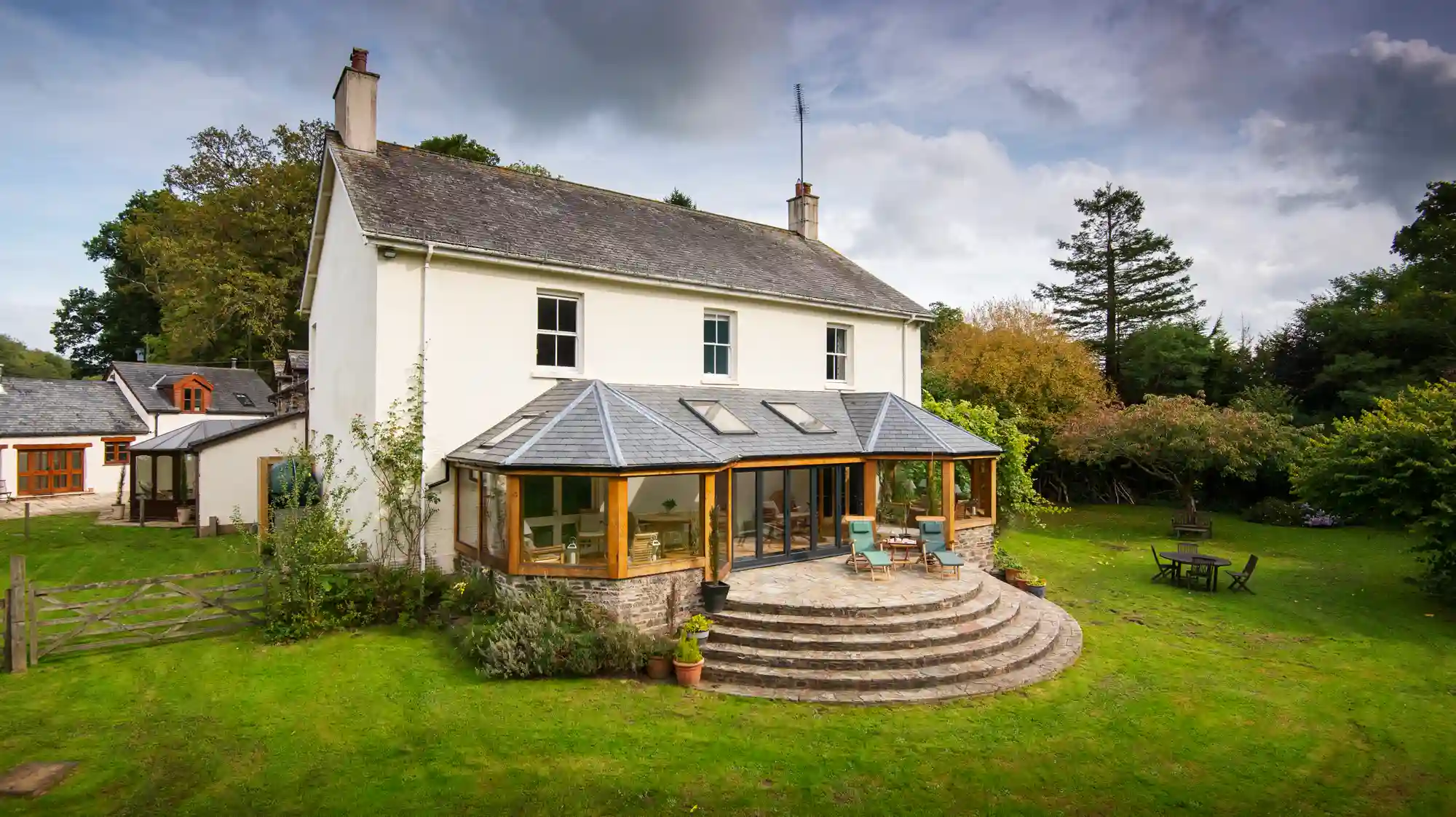 A white two-story house with a large glass conservatory and a stone patio overlooking a green lawn.