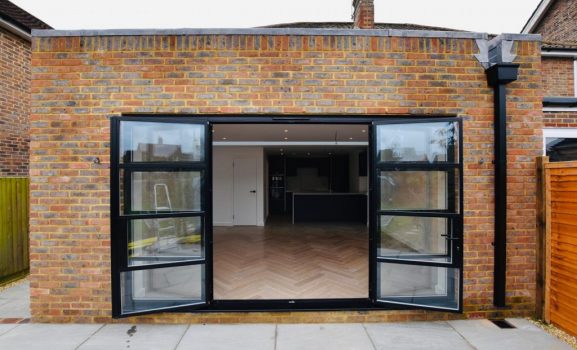 177.34-Heritage-aluminium-bifold-doors-1024×682 Brick building with open glass door entrance and wooden floor.