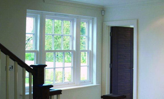 timber-windows-slide-and-tilt A bright hallway with white walls, large windows, a wooden staircase, and a dark wood door.